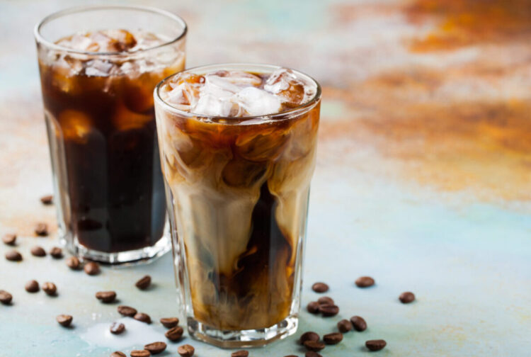 Ice coffee in a tall glass with cream poured over and coffee beans. Cold summer drink on a blue rusty background with copy space.